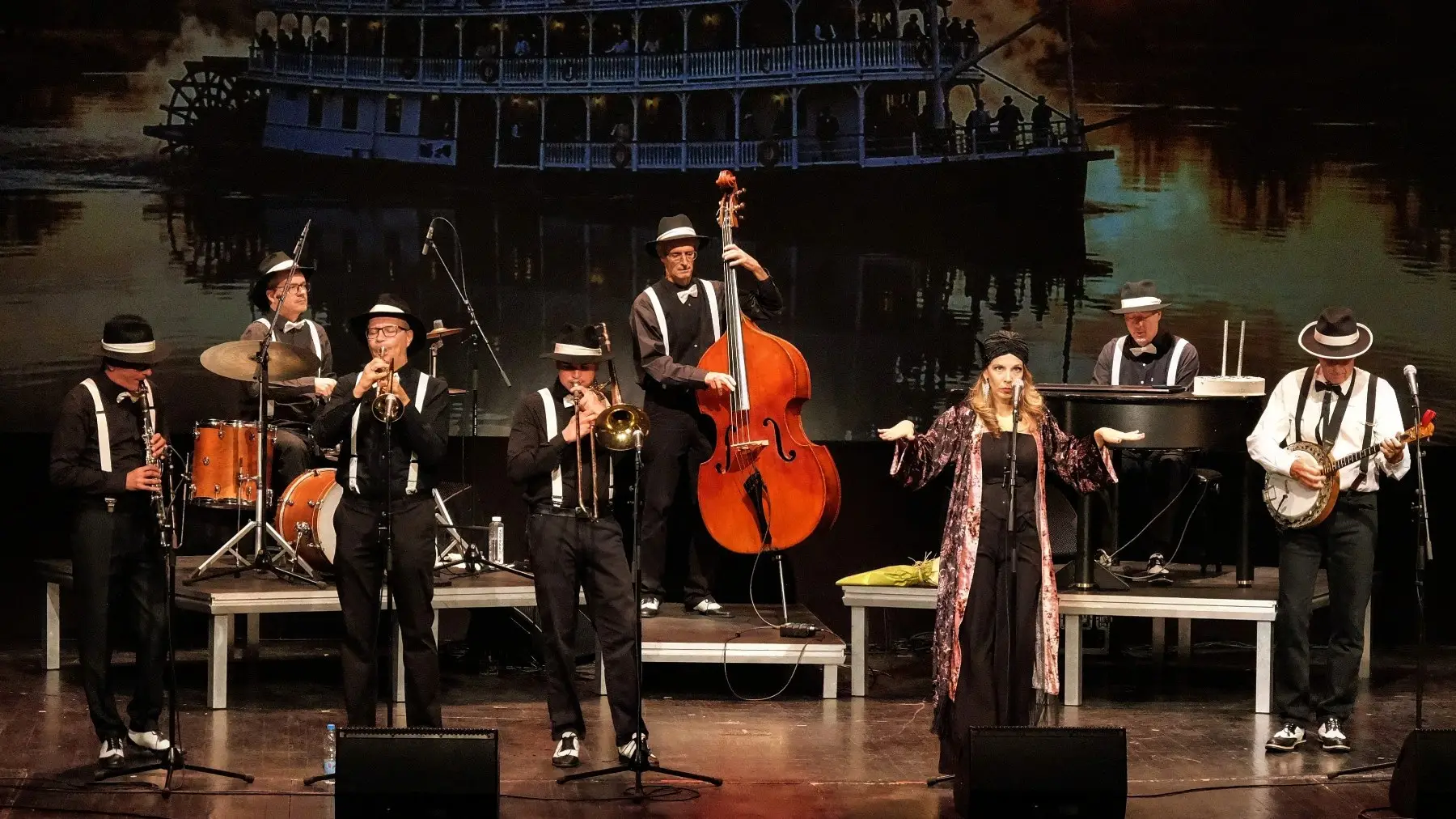 The Belgrade Dixieland Orchestra on stage in hats and braces, in front of a riverboat backdrop